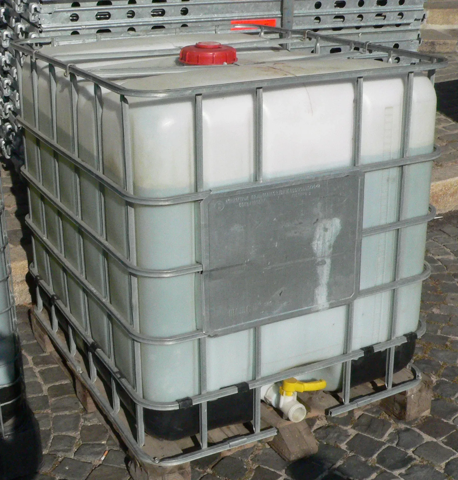 Caged plastic IBC tote on a wooden pallet with a red top fill cap and a yellow bottom discharge valve