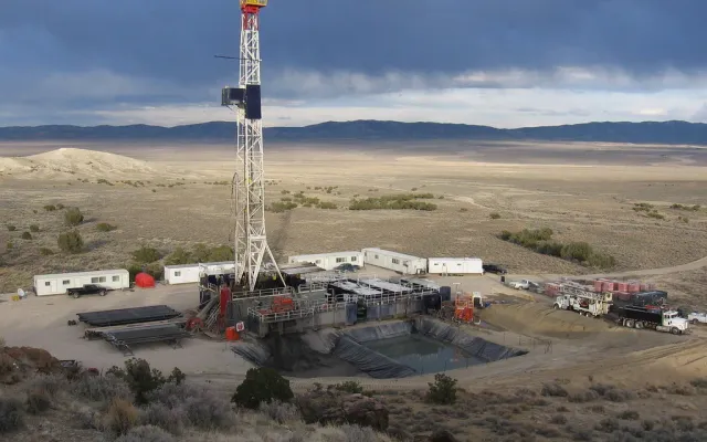 Drilling rig on a gravel well pad with pipe racks, crew trailers, trucks and a lined reserve pit in the foreground