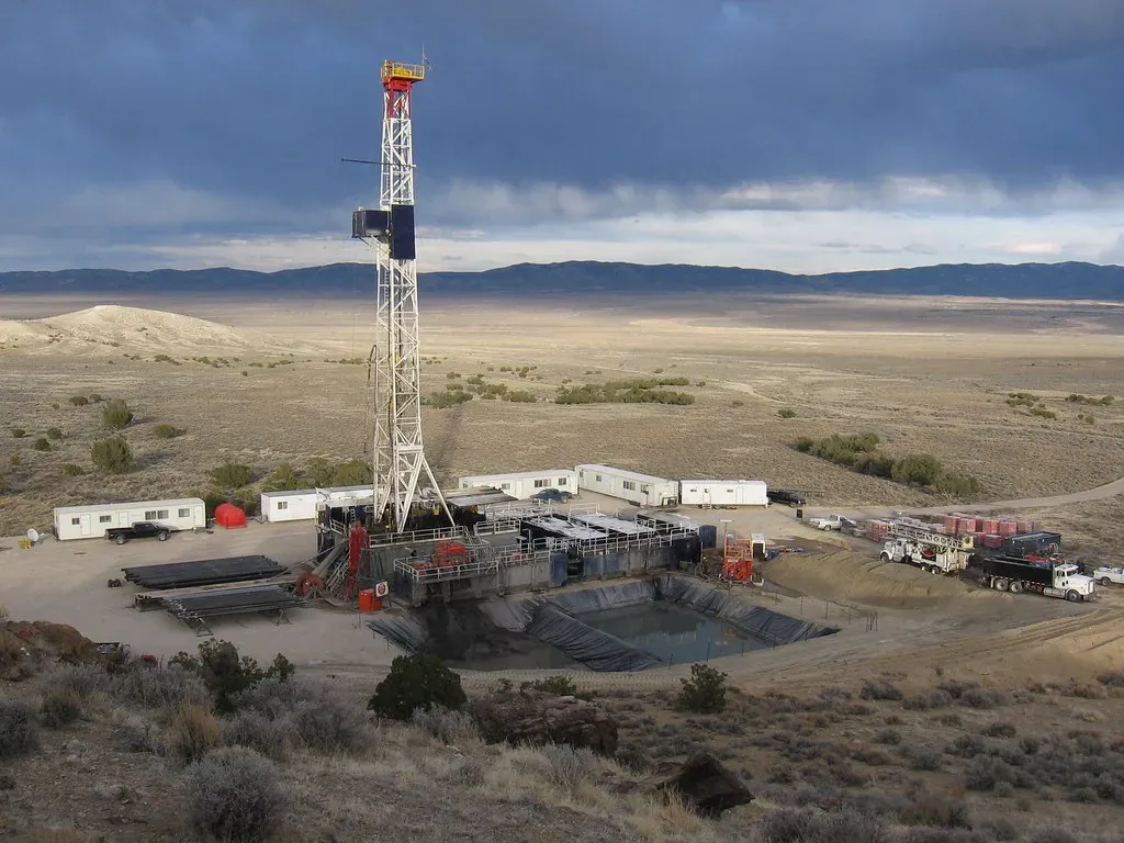 Drilling rig on a gravel well pad with pipe racks, crew trailers, trucks and a lined reserve pit in the foreground
