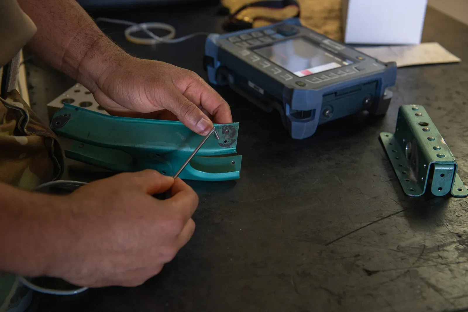 Technician's hands holding an eddy current probe against a painted metal aircraft part beside a handheld eddy current instrument