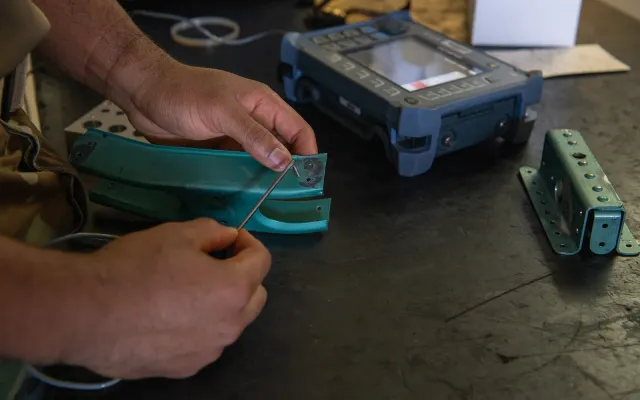 Technician's hands holding an eddy current probe against a painted metal aircraft part beside a handheld eddy current instrument