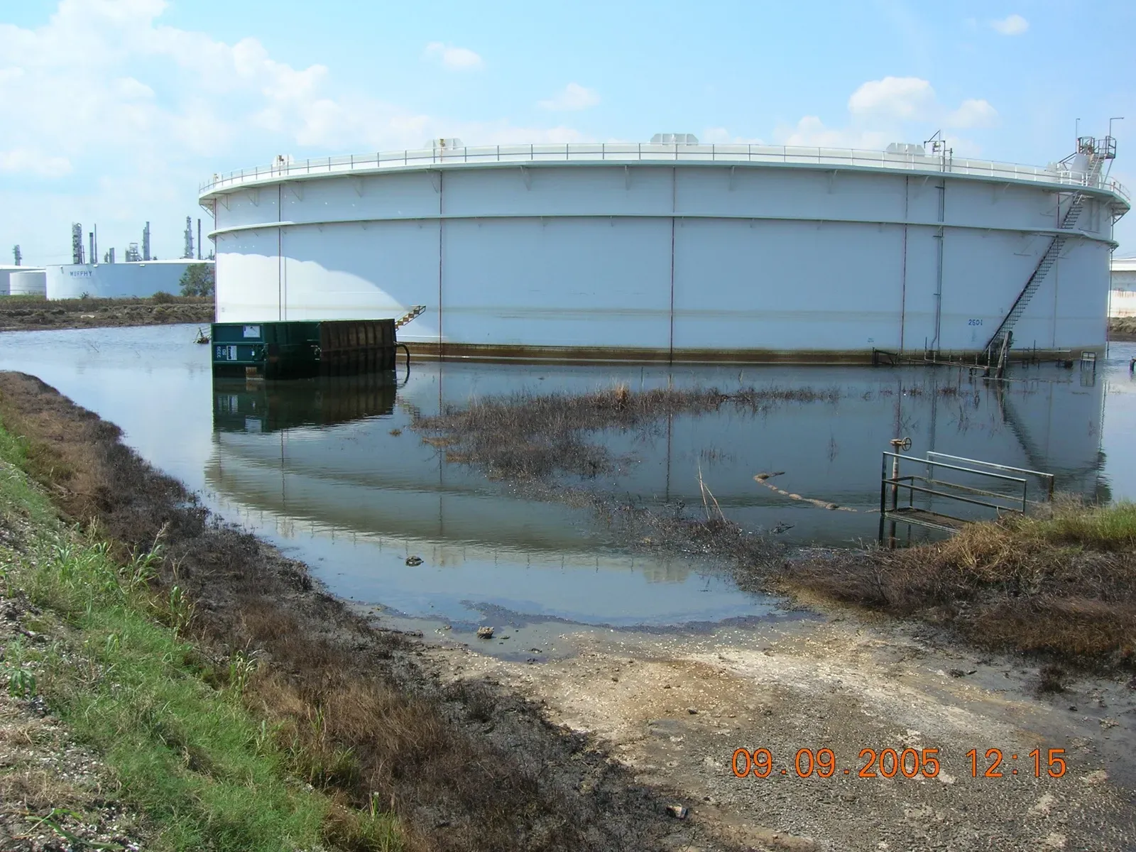 Large white storage tank standing in floodwater inside an overtopped containment berm