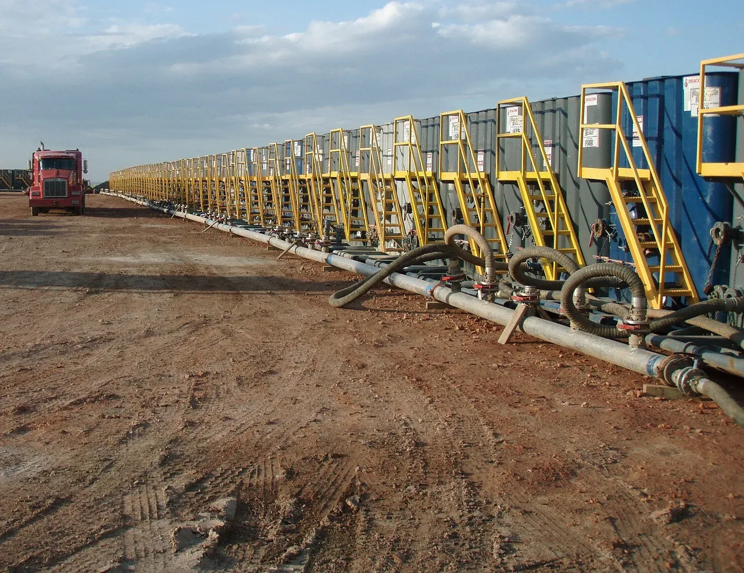 Long row of frac tanks with yellow stairways and a manifold of hoses on a dirt pad, the kind of tank line a spill containment berm goes under