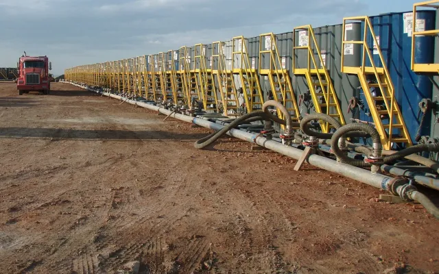 Long row of frac tanks with yellow stairways and a manifold of hoses on a dirt pad, the kind of tank line a spill containment berm goes under