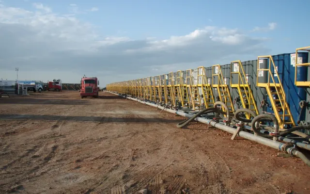 Row of frac tanks lined up on a dirt pad with hoses connected, ready for a hydraulic fracturing job