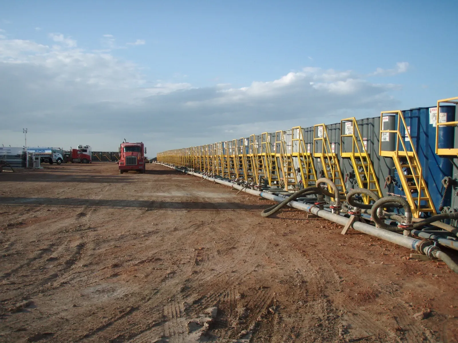Row of frac tanks lined up on a dirt pad with hoses connected, ready for a hydraulic fracturing job