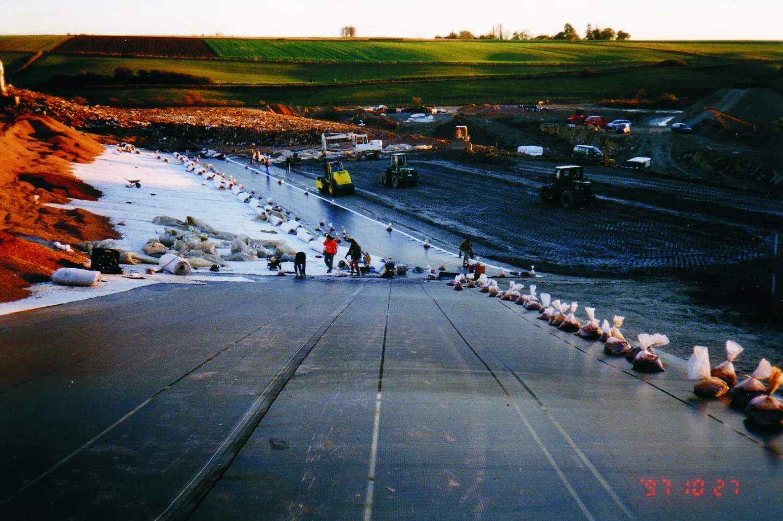 Long panels of black geomembrane liner being rolled out and weighted with sandbags on a slope during a base liner installation