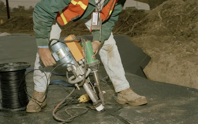 Worker in hi-vis vest using a hand-held welder to seal a containment liner geomembrane seam on a prepared subgrade