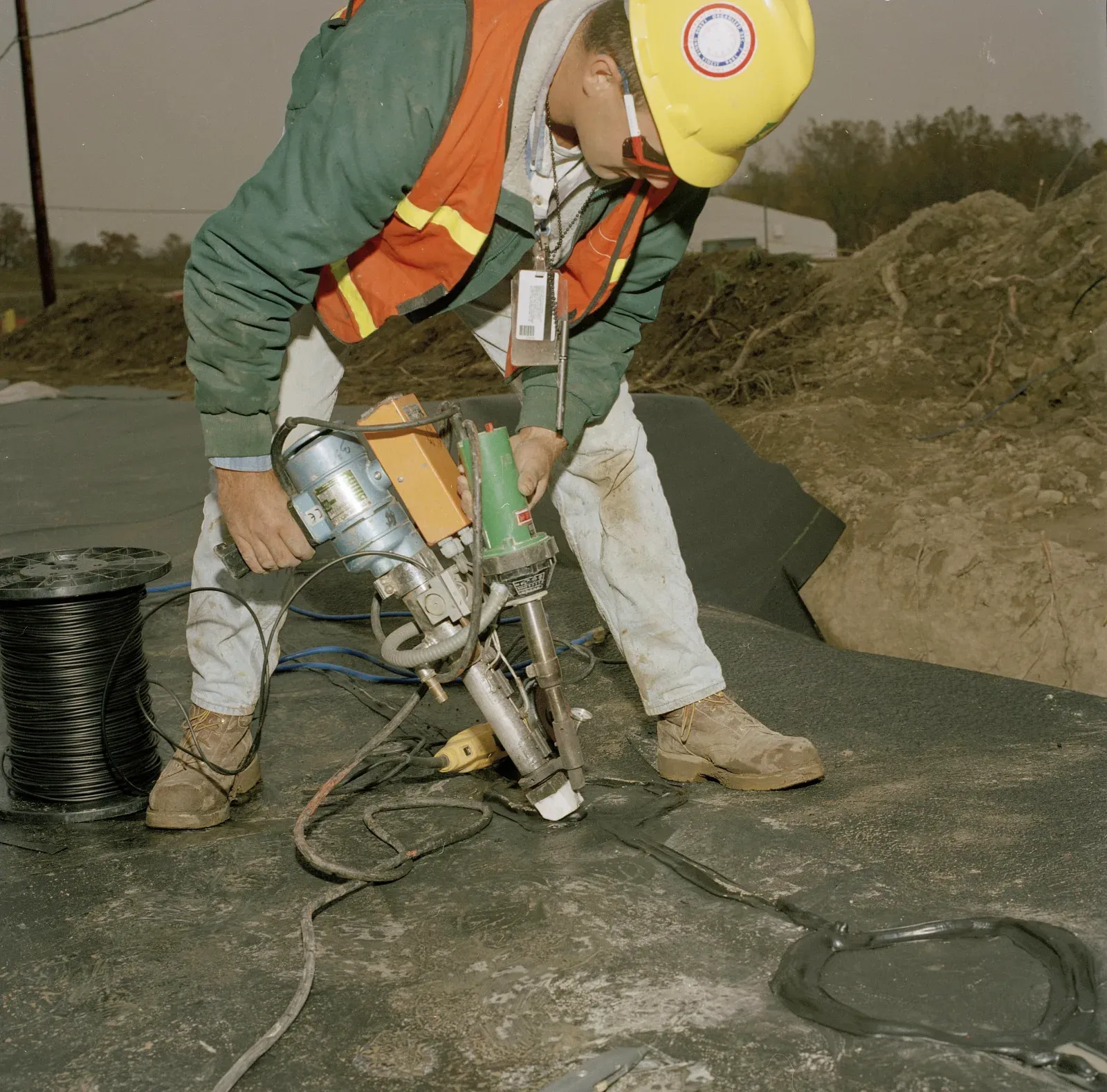 Worker in hi-vis vest using a hand-held welder to seal a containment liner geomembrane seam on a prepared subgrade
