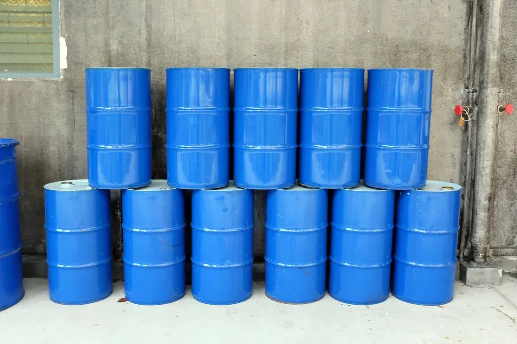Blue steel 55 gallon oil drums stacked two high on bare concrete against a wall with no spill containment under them