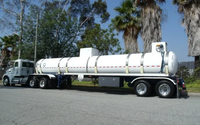 Stainless tank trailer behind a truck tractor, the kind of oil patch tanker that parks inside a drive-over containment berm to load or unload