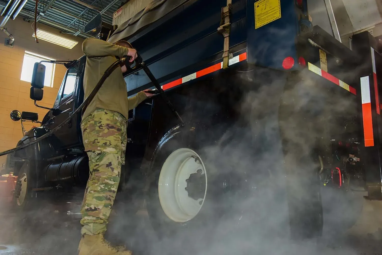 Worker pressure washing the side of a dump truck inside a wash bay, with fog from the hot water filling the lower half of the bay