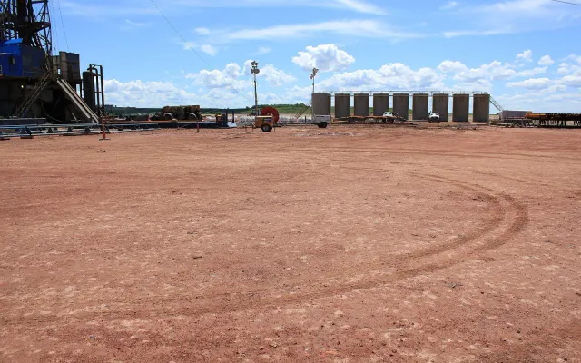 Tank battery at the far edge of a large graded dirt pad with a drilling rig on the left
