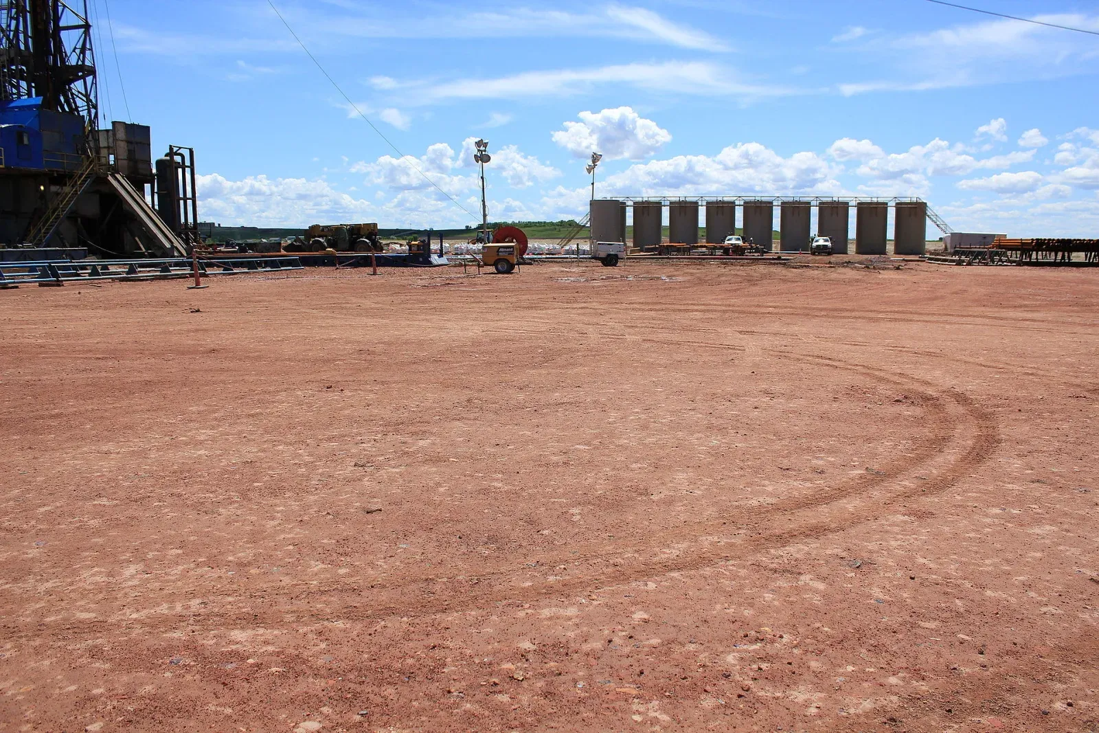 Tank battery at the far edge of a large graded dirt pad with a drilling rig on the left
