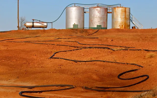 Three weathered oil tanks and a horizontal vessel on a rise of bare red earth with flowlines snaking across the ground on a Texas lease
