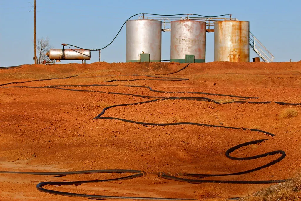 Three weathered oil tanks and a horizontal vessel on a rise of bare red earth with flowlines snaking across the ground on a Texas lease