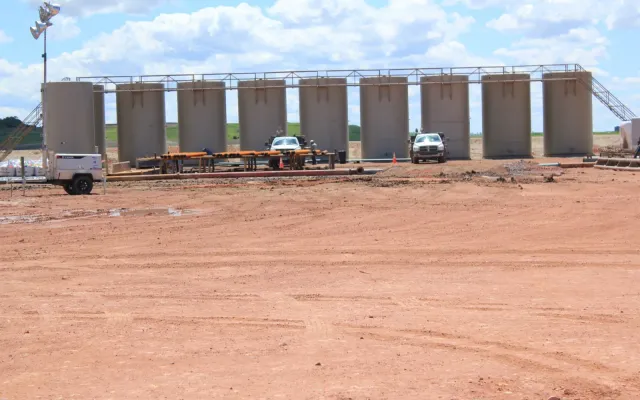 Row of tan oilfield storage tanks in a tank battery on a cleared dirt pad